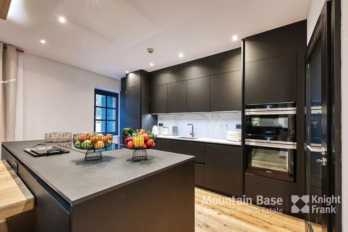 A sleek, dark modern kitchen with a large island topped with fruit bowls, black cabinets, integrated ovens, white marble backsplash, and wooden floors.