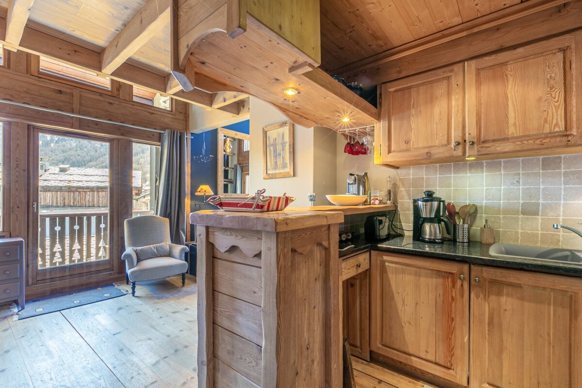 A rustic wooden chalet interior features a compact kitchen, a cozy armchair by a balcony with a mountain village view. Warm and inviting aesthetic.
