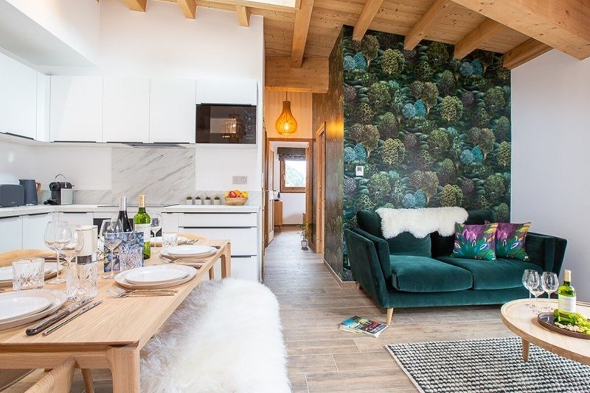Modern open-plan living room and kitchen featuring a set dining table, green velvet sofa, and forest-themed accent wallpaper.