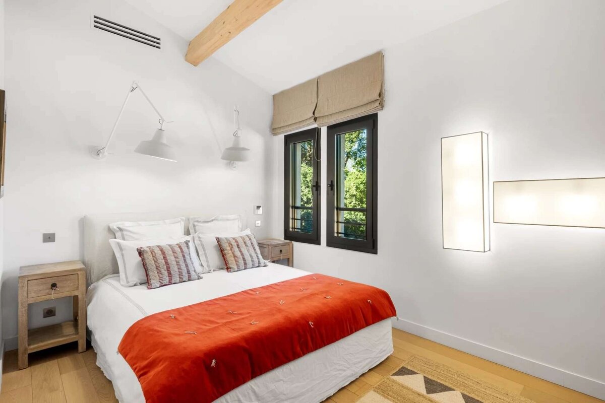 A minimalist bedroom with a white bed, vibrant orange bedspread, wooden beam, and large window overlooking lush greenery. Light wood floors and modern lighting.