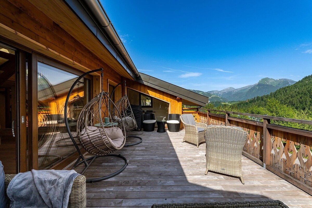 Sunny wooden balcony with hanging chairs and wicker furniture, offering stunning views of green mountains under a clear blue sky.