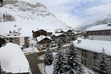 A snow-covered mountain village with traditional buildings and evergreen trees, nestled against a backdrop of vast, snowy peaks under a cloudy sky.