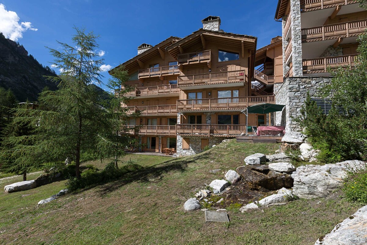 A large, multi-story wooden chalet with many balconies sits on a grassy slope under a clear blue sky, surrounded by trees and mountains.