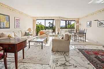 A spacious living and dining room with marble floors, white sofas, artwork, and large windows opening to a lush garden view.
