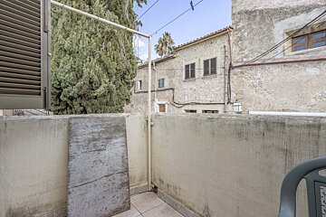 View from a small balcony with a clothesline, overlooking old stone buildings, a large cypress tree, and a distant palm under a clear sky.