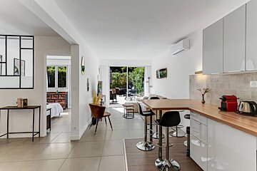 Bright, modern open-plan apartment featuring a kitchen with bar stools, a living area, and sliding doors opening to a lush outdoor patio.