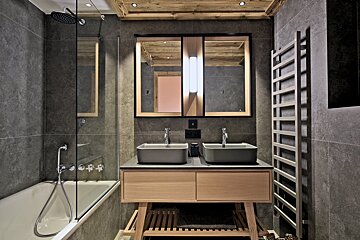 A sleek, modern bathroom featuring grey tiles, a wooden ceiling, dual vessel sinks on a light wood vanity, a bathtub with a glass screen, and a towel warmer.