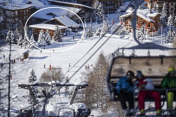 Ski resort view from a chairlift, with skiers on a snowy slope and a wooden chalet circled in white in the background.