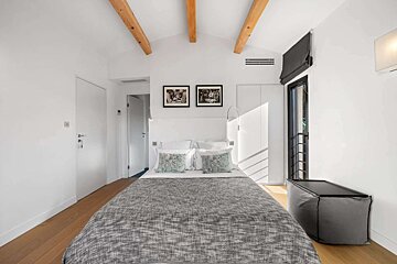 Bright, modern bedroom with a double bed, light wooden floors, white walls, exposed ceiling beams, and minimalist decor.