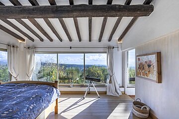 A scenic bedroom with a wall of windows revealing a lush landscape. It features exposed ceiling beams, a telescope, and a bed with blue covers.