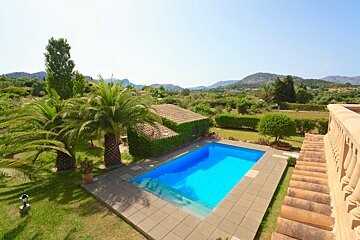 A sunny outdoor scene features a bright blue swimming pool, lush green grass, palm trees, and an ivy-covered building, all set against scenic rolling hills.