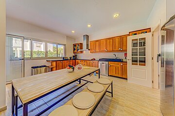 A large, bright kitchen with warm wooden cabinets, a long island/dining table with benches, stainless steel appliances, and outdoor views from windows.