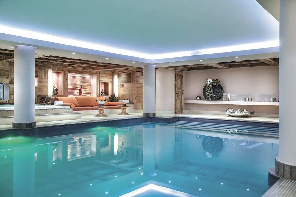 A luxurious indoor swimming pool area featuring a cozy lounge with orange seating, rustic wood paneling, and soft, ambient lighting for a relaxing spa atmosphere.