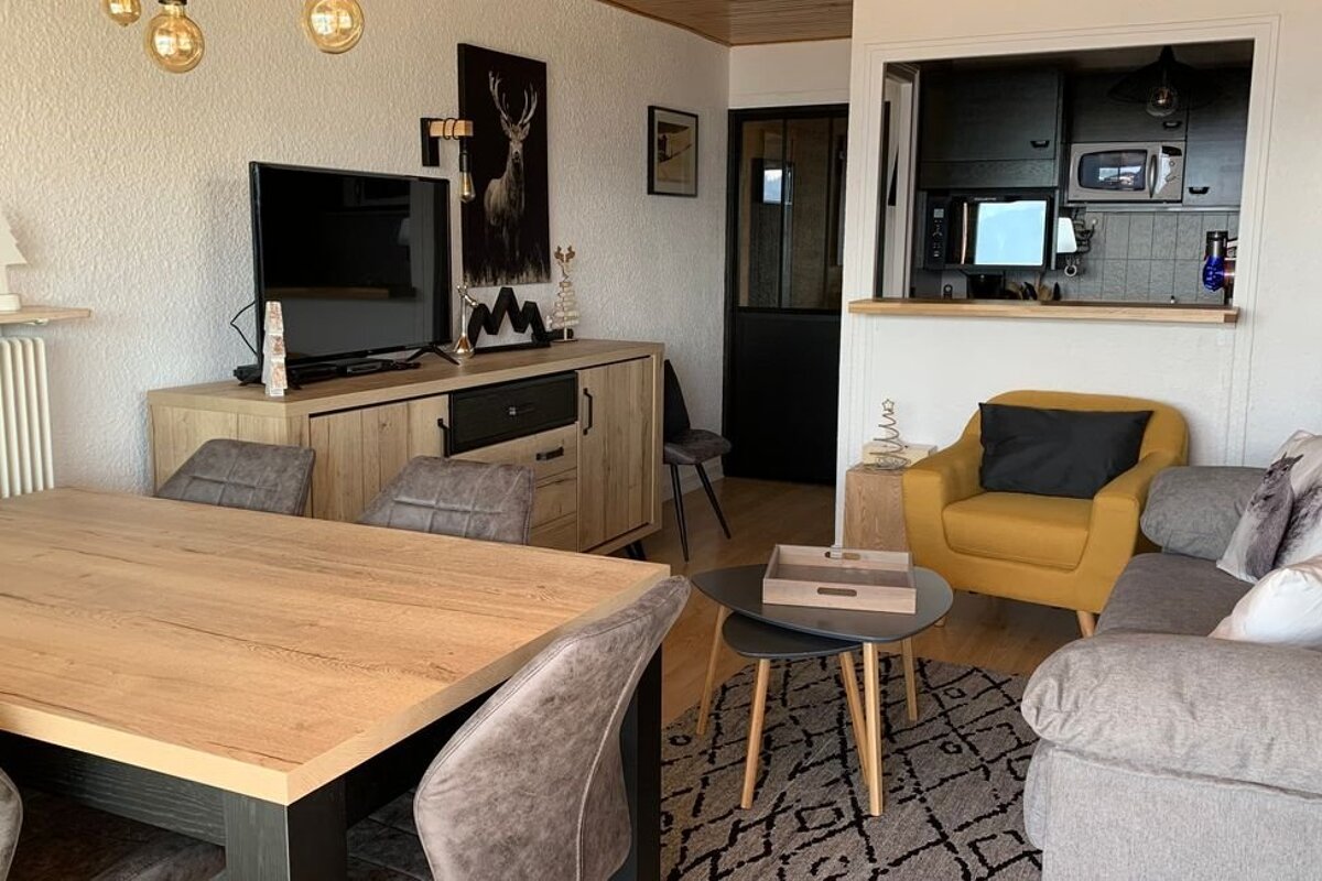 Cozy apartment interior featuring a wooden dining table, a grey sofa with a yellow armchair, and a pass-through looking into the kitchen.