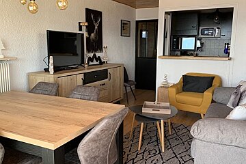 Cozy apartment interior featuring a wooden dining table, a grey sofa with a yellow armchair, and a pass-through looking into the kitchen.