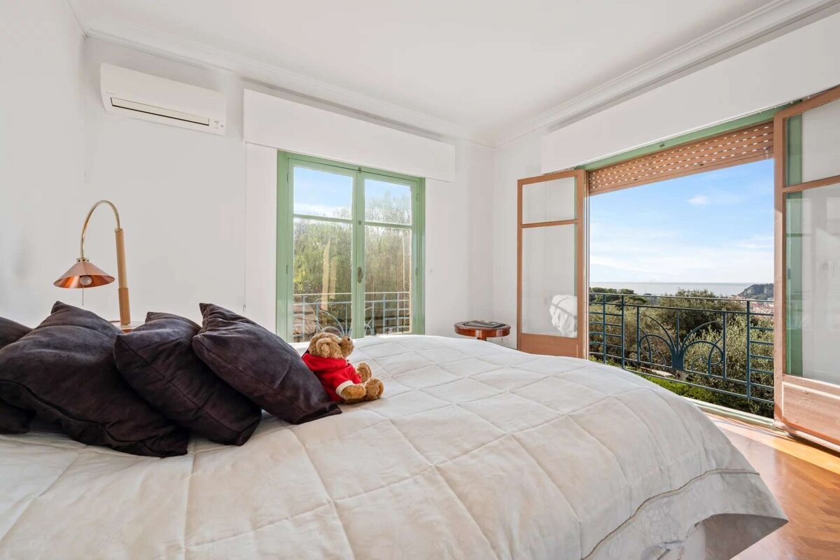 A bright bedroom with a bed, pillows, and a teddy bear, featuring open doors to a balcony with a beautiful sea view and lush greenery.