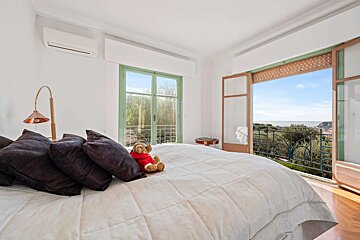 A bright bedroom with a bed, pillows, and a teddy bear, featuring open doors to a balcony with a beautiful sea view and lush greenery.