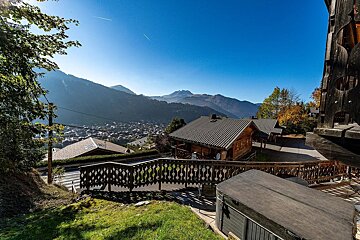Sunny view of a mountain village with wooden chalets and distant peaks under a clear blue sky. A hot tub and decorative wooden railing are in the foreground.