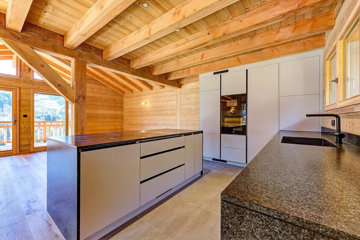 Modern chalet kitchen featuring sleek white cabinets, dark stone countertops, and rustic exposed wooden beams.