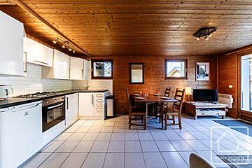 A cozy, wood-paneled room features a modern white kitchen, a wooden dining table, and white tiled flooring, lit by windows.