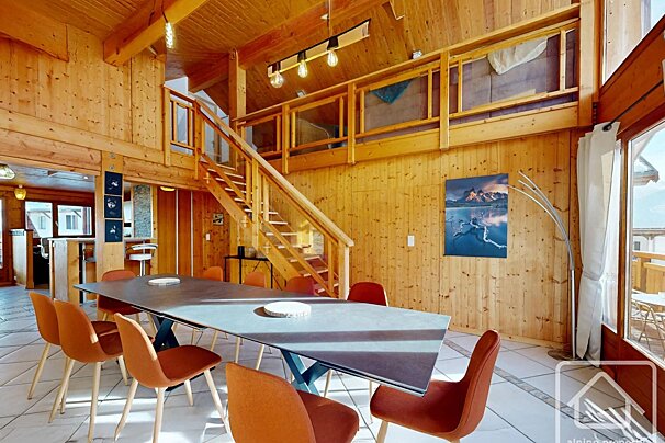 Rustic wooden chalet interior with a long dining table, orange chairs, and a wooden staircase leading up to a loft.