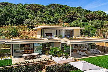 Modern luxury villa with large glass walls and green roofs, set against a lush, forested hillside.