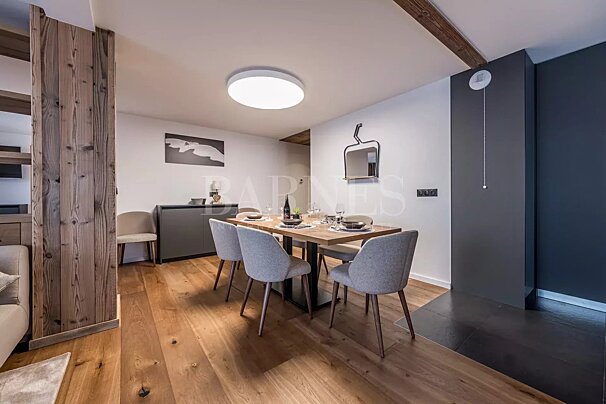 A modern dining area with a wooden table, grey chairs, and light wood flooring. Rustic accents include a wooden pillar and exposed beam, with a ski lift mirror.
