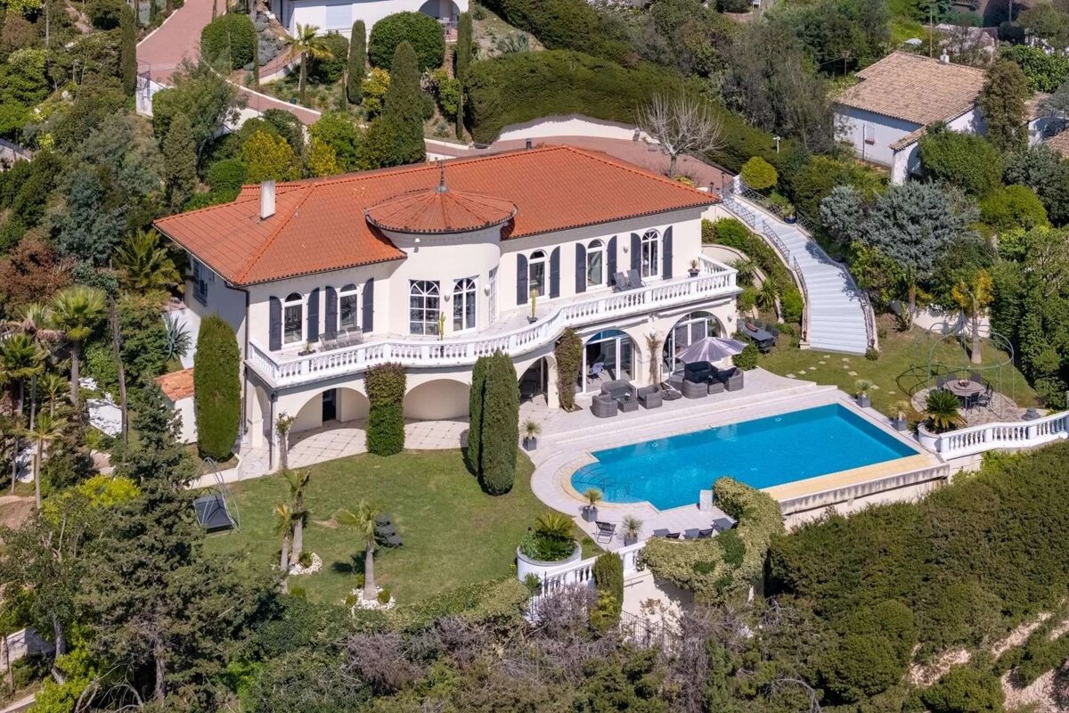 Aerial view of a grand villa with a red roof, a large blue swimming pool, and lush green gardens, situated on a hillside.