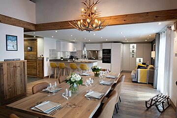 Spacious, modern open-plan room with dining, kitchen, and living areas. Features a striking antler chandelier, rustic wood beams, and yellow accents.