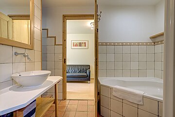 A tiled bathroom features a white vessel sink and corner tub. An open wooden doorway reveals an adjacent room with a black couch and wall art.