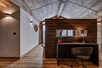 A modern chalet interior featuring a vaulted wooden ceiling, light flooring, and a dark wood desk with a window view of a snowy forest.