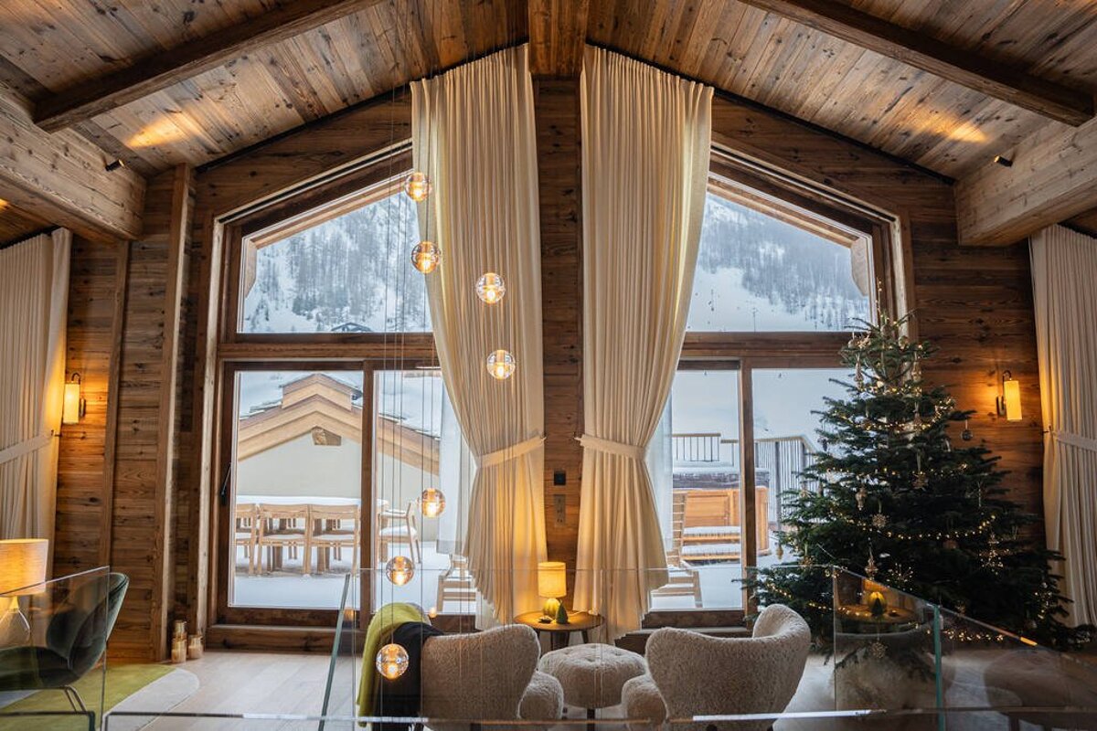 Warm wooden ski chalet with large windows overlooking snowy mountains. Features a decorated Christmas tree and modern lighting, creating a cozy winter scene.