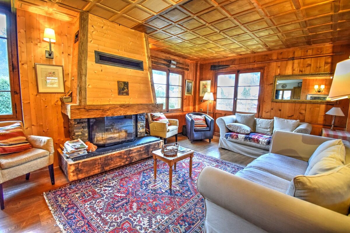 A warm, wood-paneled living room featuring a grand stone fireplace, plush seating, and an intricate red rug. Cozy and inviting.