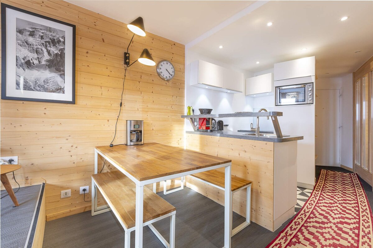 A modern, wood-paneled kitchen and dining area with white cabinets, a table and benches, mountain art, and a red patterned rug in the hallway.