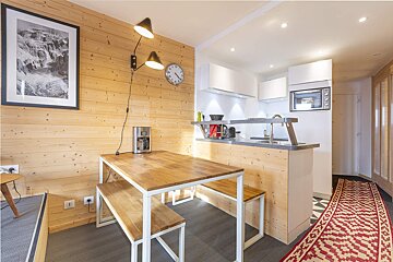 A modern, wood-paneled kitchen and dining area with white cabinets, a table and benches, mountain art, and a red patterned rug in the hallway.