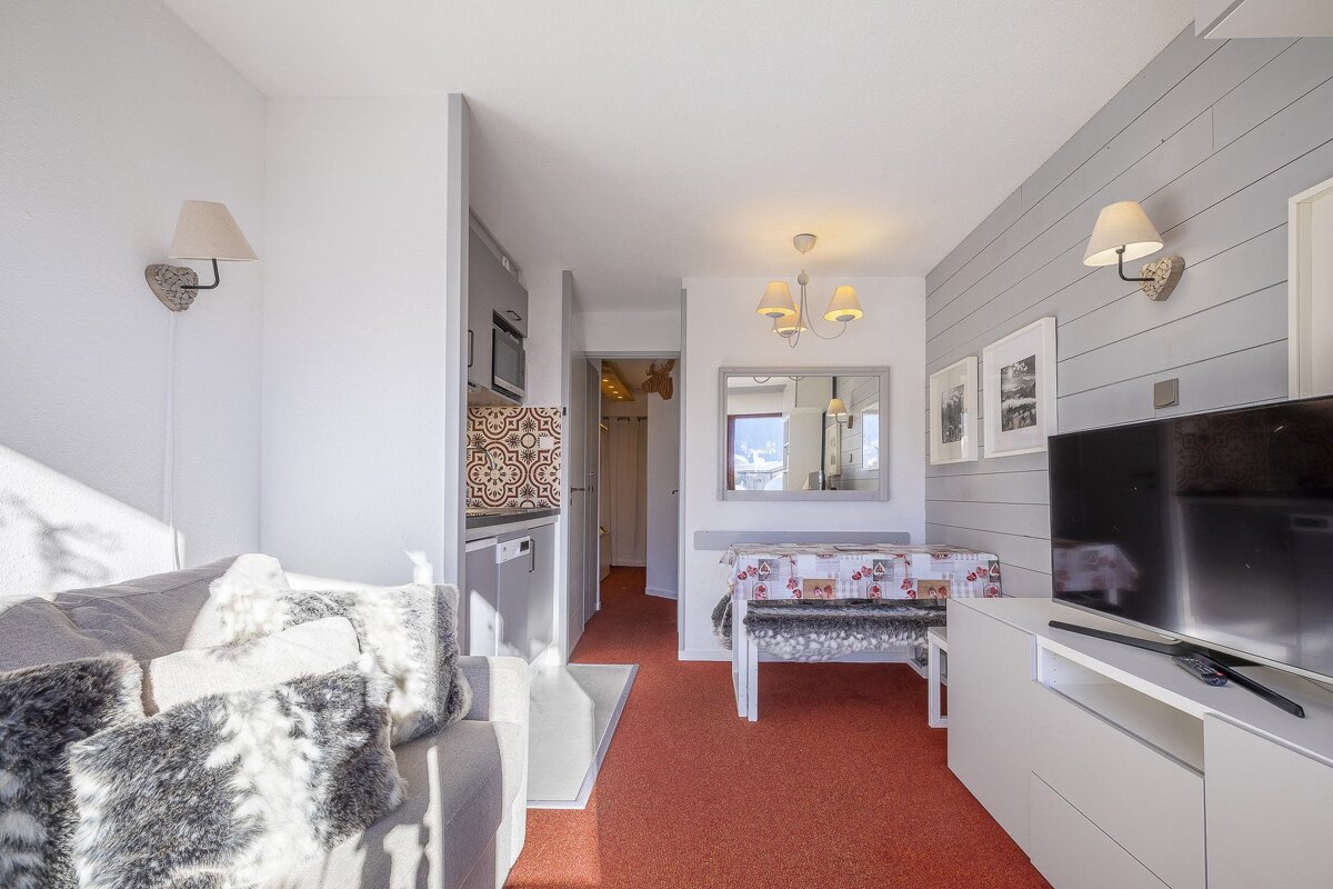 Cozy, modern studio apartment with a gray couch, small kitchen, dining table, TV area with a shiplap wall, and vibrant red carpet.