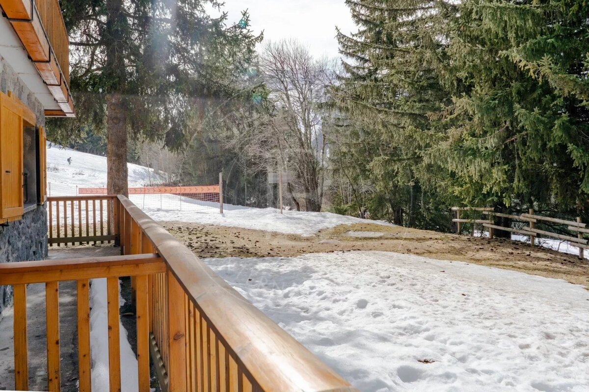 A view of a snowy ski resort, as seen from a wooden balcony with pine trees on a sunny winter day.