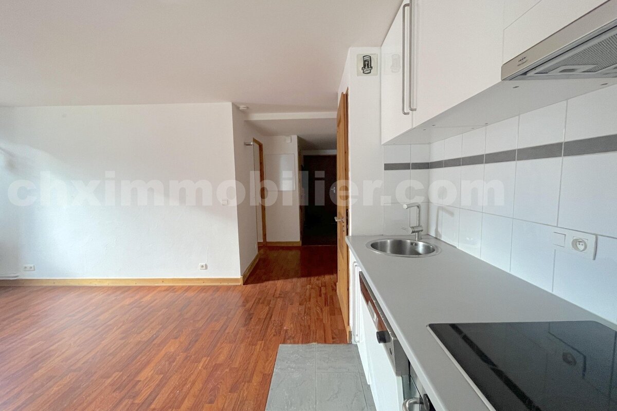 Modern open-plan studio apartment featuring a compact white kitchen with a sink and stovetop, and an adjacent living area with wooden flooring.