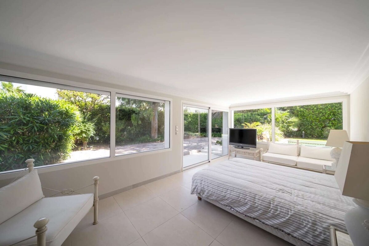 Bright bedroom with large windows/glass walls overlooking a lush garden and patio area. Features a bed, sofa, and TV.