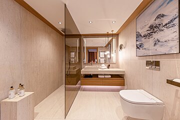 Modern bathroom with beige stone tiles, a glass walk-in shower, floating vanity, white toilet, and mountain artwork.