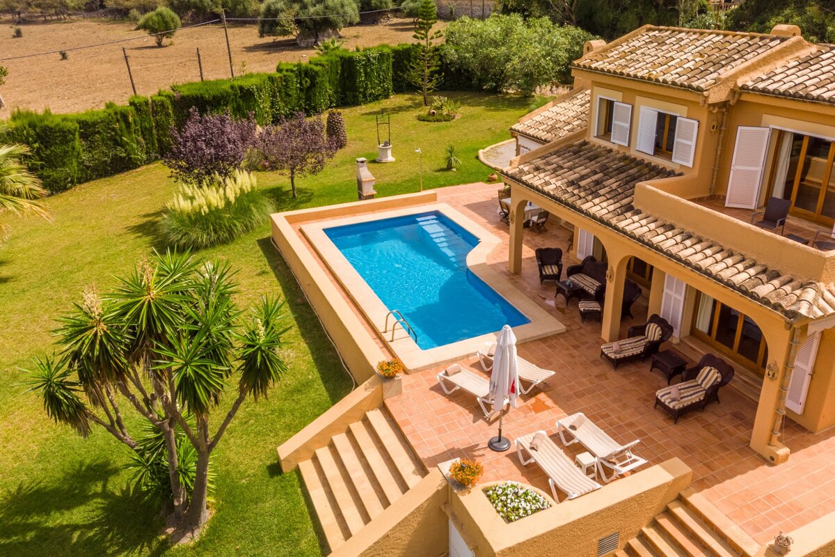 Sunny aerial shot of a spacious villa with a blue pool, tiled patio, and extensive green lawn, surrounded by lush vegetation.