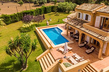 Sunny aerial shot of a spacious villa with a blue pool, tiled patio, and extensive green lawn, surrounded by lush vegetation.