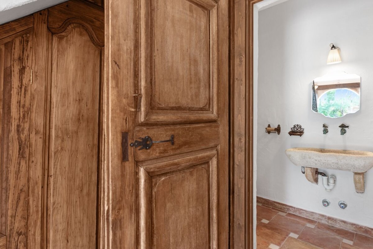A rustic wooden door with an old latch partially opens to a white bathroom featuring a carved stone trough sink, mirror, and terracotta floor.