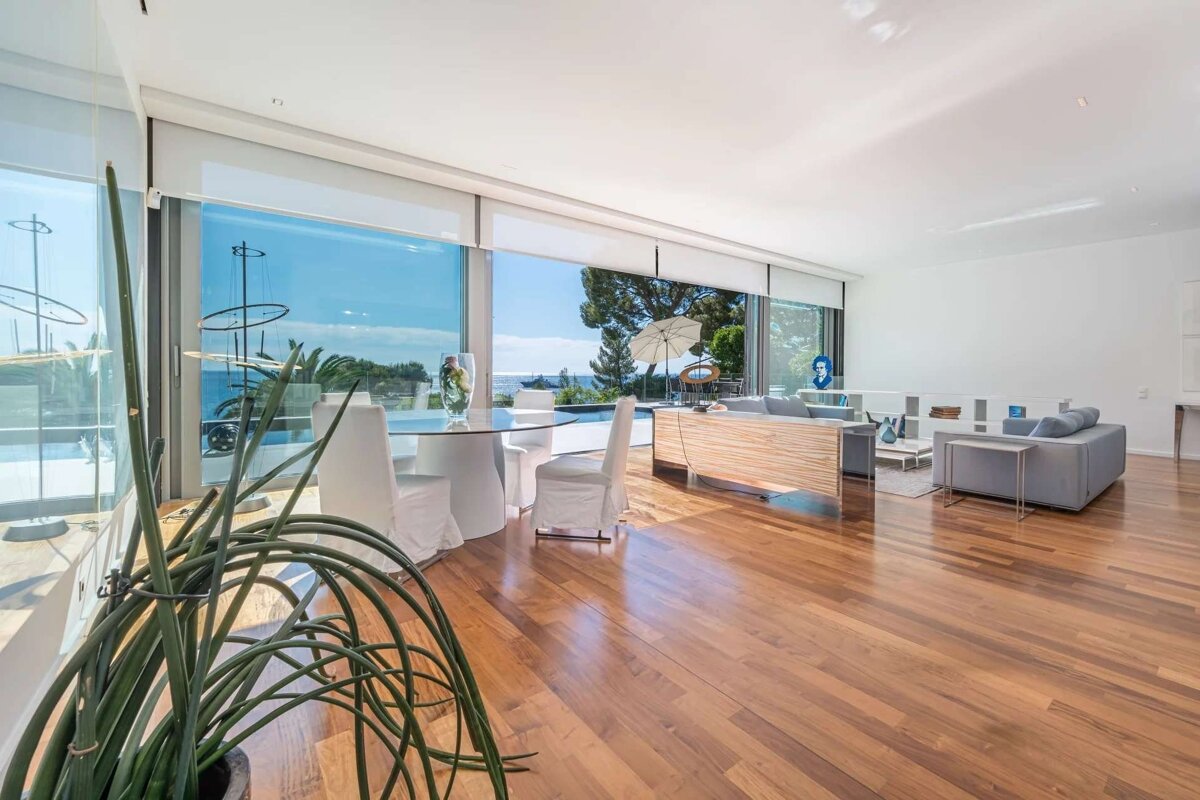 Spacious, modern living room with floor-to-ceiling windows offering a stunning sea view. Features a dining area, lounge, and warm wooden floors.