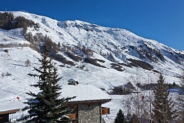 Sunny snow-covered mountains with ski tracks, a stone house, and pine trees beneath a clear blue sky.