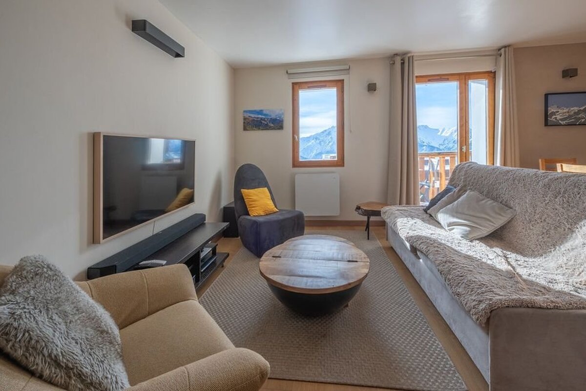 Cozy living room with comfortable seating, a round wooden coffee table, TV, and a window opening to snowy mountain views.