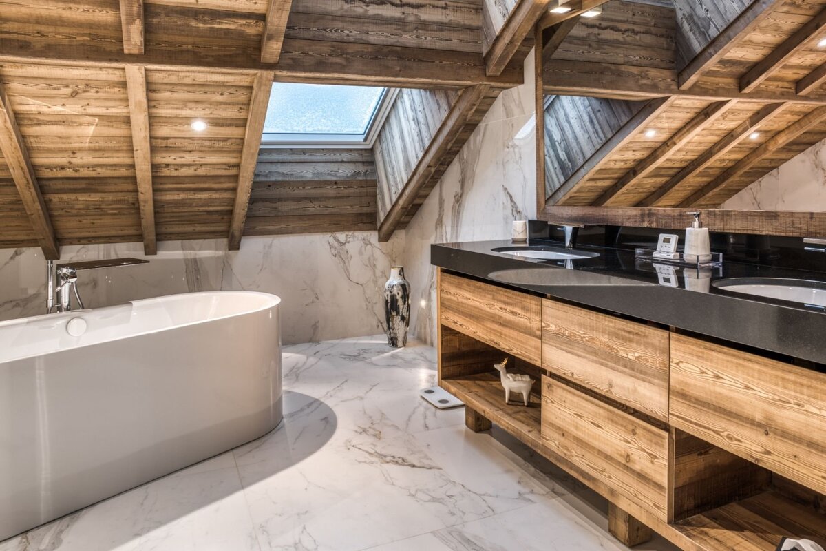 A rustic-modern attic bathroom featuring wooden beams, marble walls/floor, a freestanding tub, double vanity, and a bright skylight.