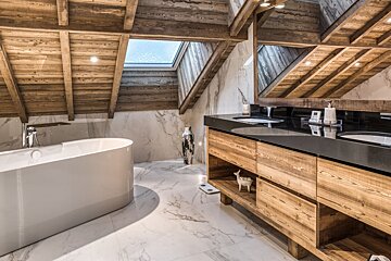 A rustic-modern attic bathroom featuring wooden beams, marble walls/floor, a freestanding tub, double vanity, and a bright skylight.