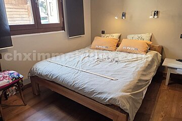 A cozy bedroom features a wooden bed with a light duvet, deer pattern, and warm orange pillows. Wall lamps provide light near a window with dark blinds and wooden floors.