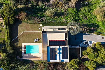 Aerial shot of a house featuring a swimming pool, solar panels on the roof, a car on the drive, and lush green surroundings.